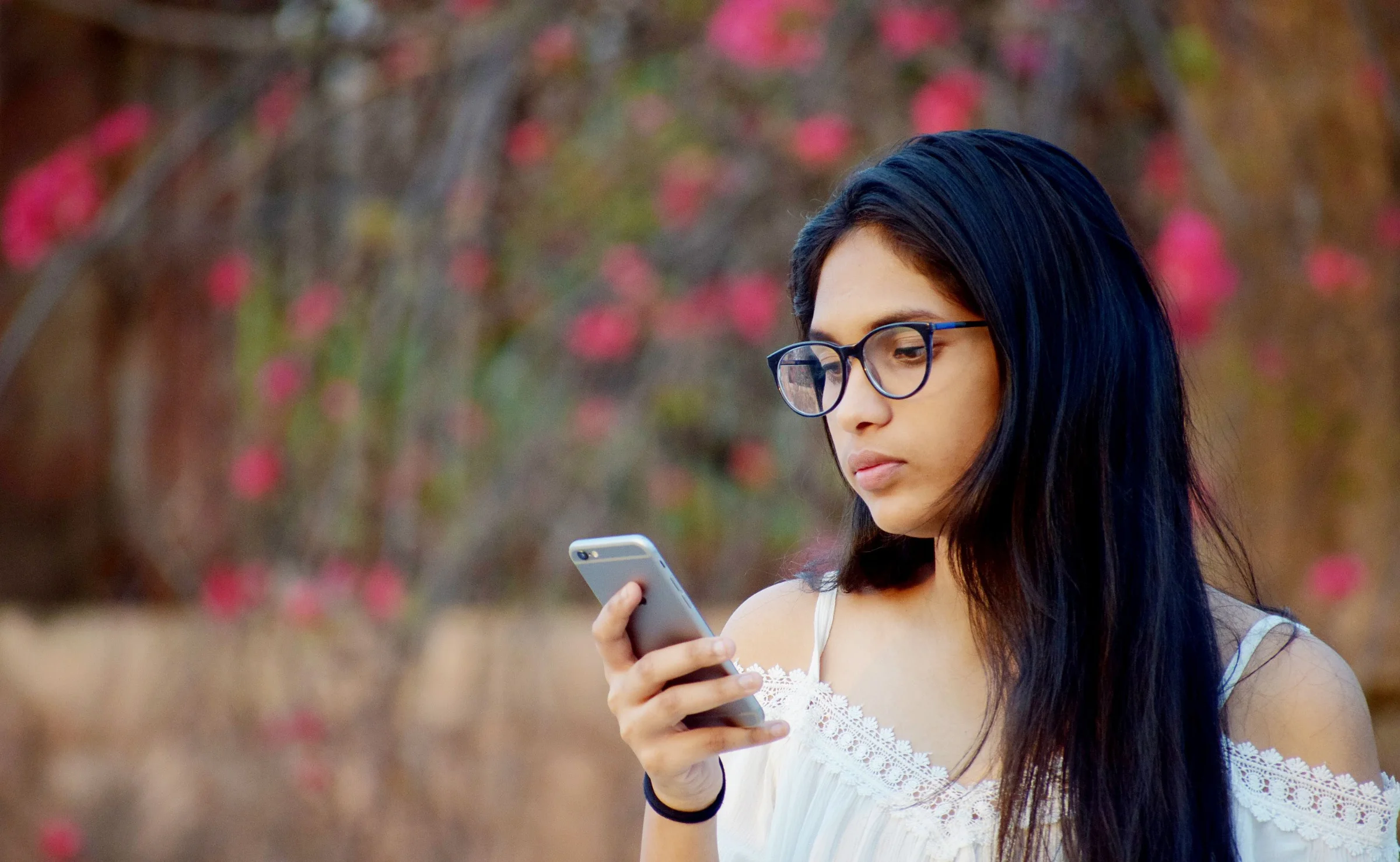 A young woman in glasses reads a question on her phone outdoors, pink blossom behind her